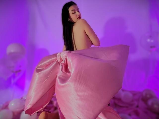 IrisBlair with long black hair, naked, covered by a pink satin sheet, back turned, looking over shoulder, purple and pink lighting, soft balloons in the background, sexy pose.