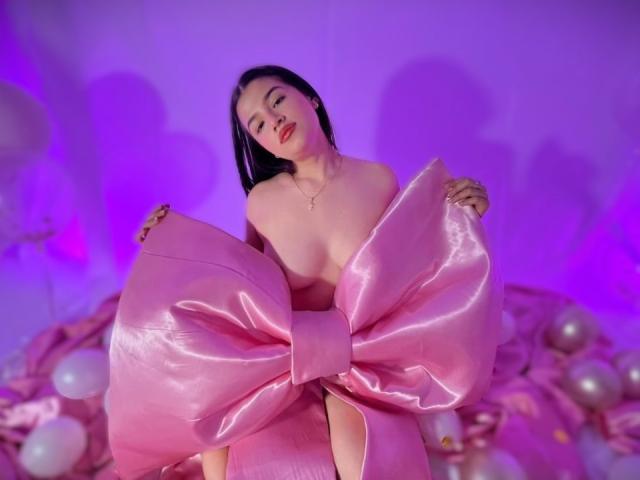 Iris Blair, nue, tenant un énorme nœud en satin rose, exposant ses seins. Éclairage violet et bleu. Ballons roses et pétales sur le sol. Tension sexuelle, pose provocante, expression séduisante..