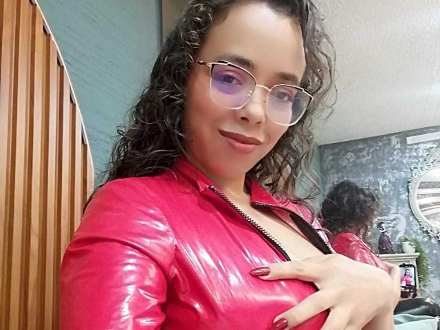 Barbara Leythonn, cheveux noirs et bouclés, portant une veste rouge brillante en latex, de grandes lunettes transparentes, un sourire en coin, une main sur la poitrine, décolleté visible, rouge à lèvres rouge vif, intérieur, chaise en bois, mur gris, reflet dans la fenêtre.