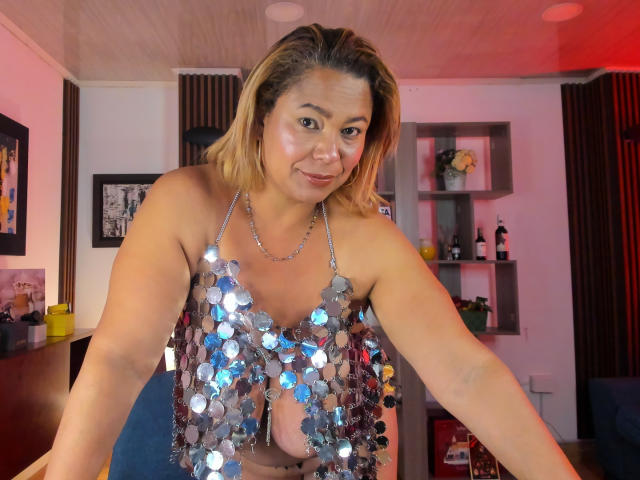 VanesaStella, inclinada para frente, vestindo uma blusa prateada com lantejoulas que revela seus seios, com um sorriso sedutor. Ela tem a pele de cor clara, castanho, e cabelo loiro que chega aos ombros. Há iluminação vermelha e rosa ao fundo..
