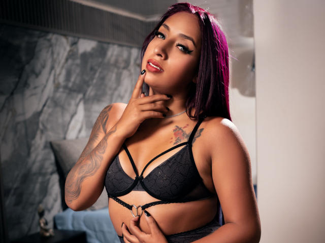 Fotografia di Roxanne Marquez, una donna attraente con capelli viola scuro, che indossa un reggiseno di pizzo nero e tocca il suo dito. sulle sue labbra, tatuaggi sul braccio destro, sguardo seducente, illuminazione soffusa.