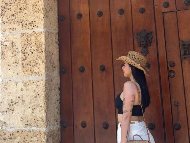 IsabellaJames, with black hair and tattoos, stands against a wooden door wearing a straw hat, black top, and white pants. Sunlight casts shadows on her left side. Fucking sexy.