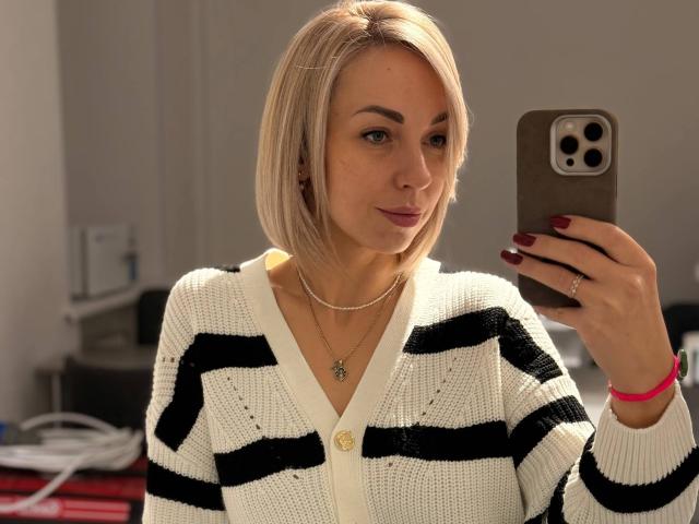 RoseGloss, una donna bionda con un taglio di capelli a caschetto, si fa un selfie con uno smartphone tenuto nella mano destra. Indossa un cardigan bianco con righe nere, una collana e un braccialetto rosso. La luce del sole illumina il suo viso.