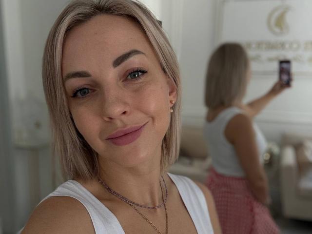 Close-up photo of RoseGloss, a blonde woman with blue eyes and pink lipstick, smiling at the camera. She's wearing a white tank top and red checkered skirt. Reflection in mirror shows her holding a phone, back to camera.