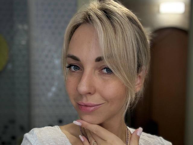 Close-up of RoseGloss, a blonde woman with light skin and blue eyes, smiling slightly, resting her chin on her hand. She's wearing a white lace top, with soft, natural lighting.