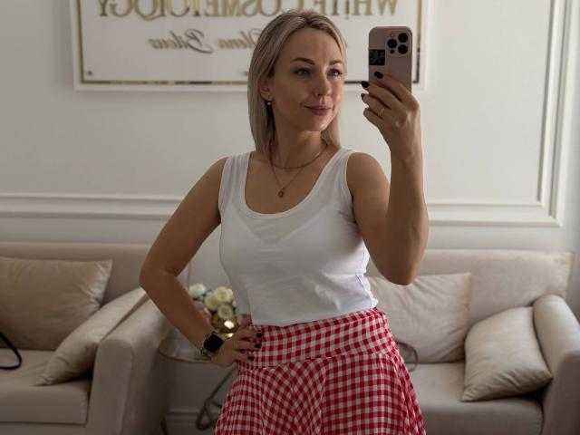 RoseGloss stands in a living room, taking a selfie with a smartphone in her right hand. She wears a white tank top and red checkered skirt, smiling subtly. Light streams in.