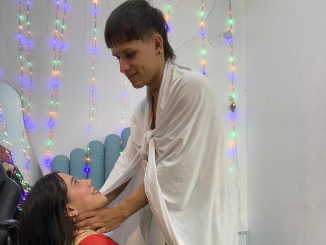 AnjaliRohan, een slanke man met kort zwart haar, die een wit shirt draagt, leunt voorover naar een brunette vrouw met lang haar. Ze draagt een rode top en houdt haar nek vast. Kleurrijke lichtslingers op de achtergrond.