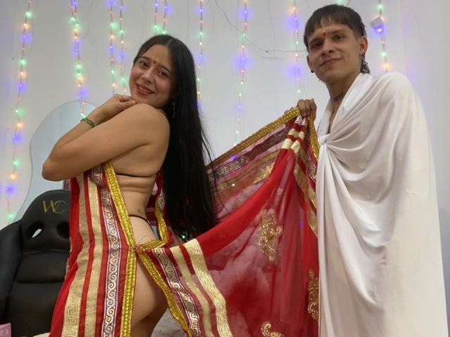 AnjaliRohan, with long black hair and medium breasts, wears a red and gold sari, exposing her back and butt. A man with short brown hair, in a white robe, holds the sari. Multicolored string lights in the background. Sexual tension, fucking, cock implied.