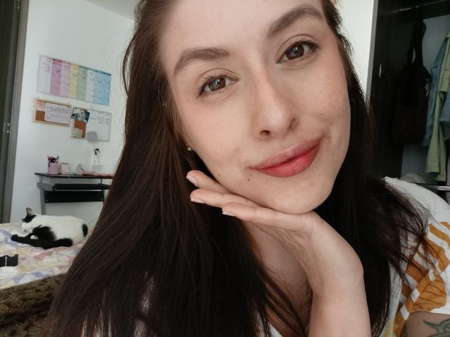 Autoritratto ravvicinato di Isabella Palacio, con la pelle chiara e i capelli castano scuro, che sorride con una mano appoggiata sulla guancia, e indossa un maglietta a righe gialle, camera da letto bianca con calendario, gatto bianco e nero sul letto. Che carino!.