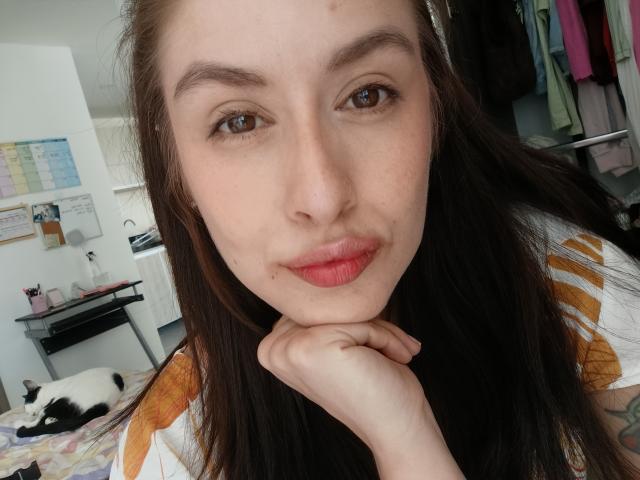 Selfie de primer plano de IsabellaPalacio, con piel pálida, ojos marrones y labios rojos, apoyando el mentón en la mano. Ella lleva una blusa blanca con estampados naranjas, y el fondo incluye un gato y una habitación desordenada..