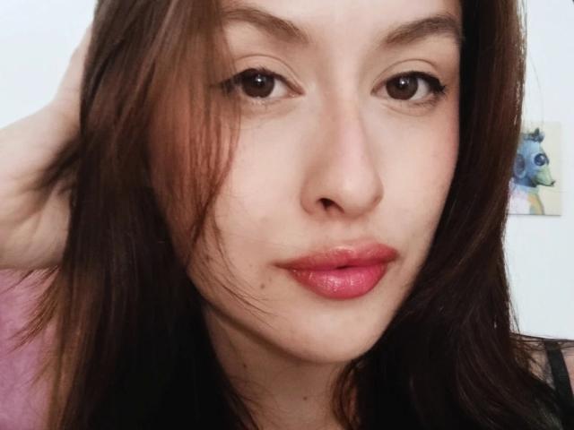 Fotografia ravvicinata di Isabella Palacio, con la pelle chiara, capelli castani, rossetto rosso e leggere lentiggini. Lei guarda direttamente la fotocamera con un'espressione leggermente seducente e rilassata. Sfondo sfocato.