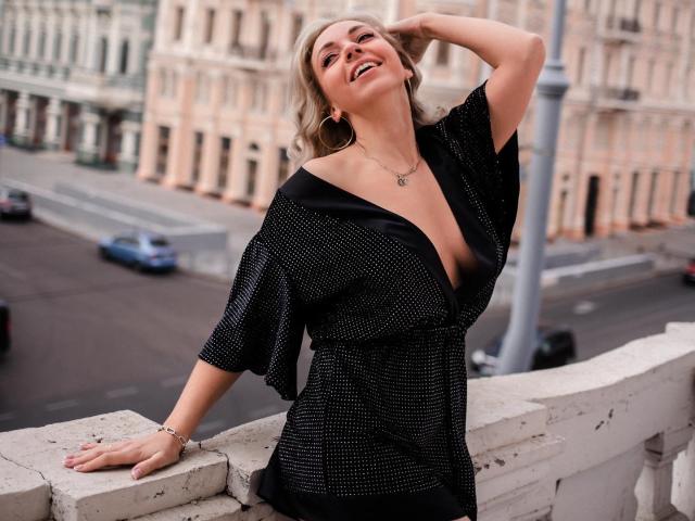 RoseGloss è in piedi su un balcone di pietra, appoggiata alla ringhiera con una mano tra i capelli biondi. Indossa un abito nero con un profondo scollo a V e piccoli pois bianchi. Sta sorridendo, mostrando un accenno di scollatura. Sfondo urbano con strada..