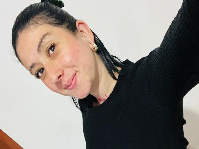 Foto em close-up de Valeriaaf, com pele clara, cabelo preto preso em um coque, vestindo uma camiseta preta texturizada, sorrindo levemente, morena. olhos, pequenas brincos, fundo branco, leve ângulo, pose casual.