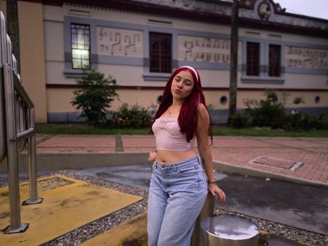 MiaLeroy, red-haired, leans against a metal pole, wearing a white crop top and high-waisted jeans, standing in an urban setting. Background shows a building with graffiti and a wet sidewalk. Fucking sexy.