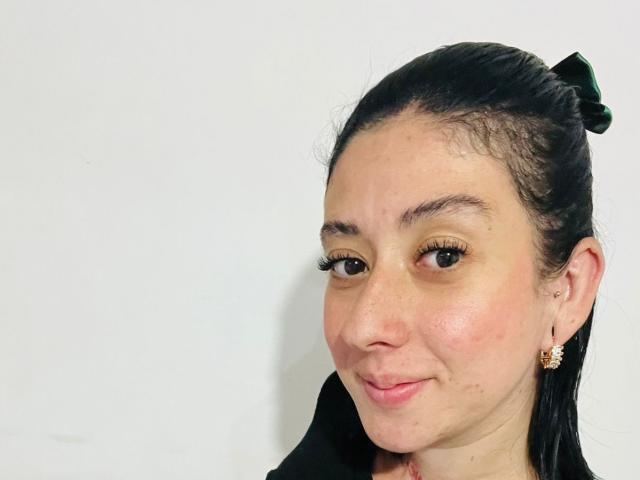Photograph of a woman, Valeriaaf, with light skin, dark hair in a bun, brown eyes, and small gold earrings. She has a slight smirk, and visible facial freckles. White background.