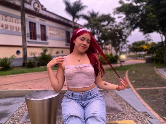 MiaLeroy, una donna dai capelli rossi che indossa un top di pizzo bianco e jeans a vita alta, è seduta su una panchina di metallo. Lei tiene in mano un sottile filo rosso, appare rilassata, con alberi e un edificio sullo sfondo..