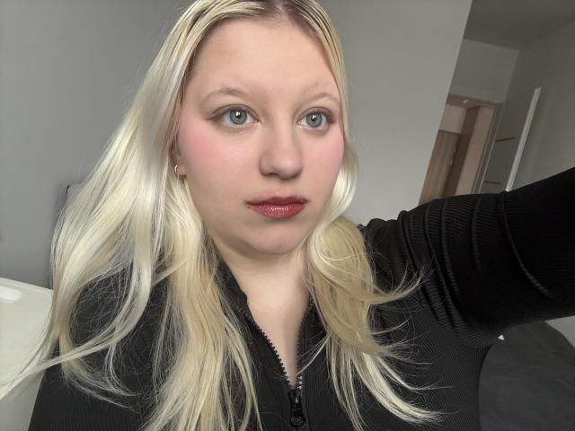 Fotografia ravvicinata di LenaHoney con la pelle chiara e i capelli biondi, che indossa una maglia nera con zip e guarda direttamente verso... fotografia di una persona con rossetto rosso, leggero rossore sulle guance, braccio esteso.