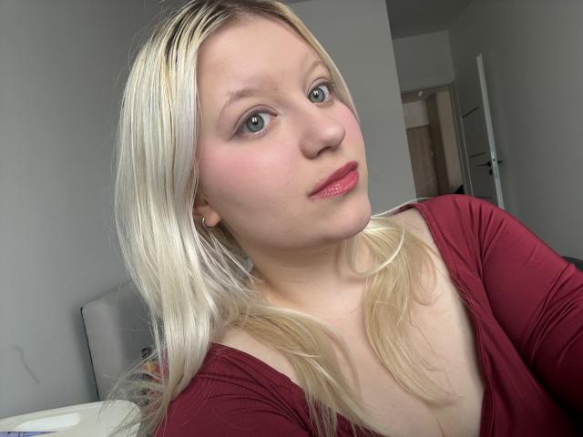 Nahaufnahme von LenaHoney mit langen blonden Haaren, blauen Augen und rotem Lippenstift, die ein burgunderfarbenes Oberteil trägt, mit leicht geneigtem Kopf. Direkter Blick in die Kamera, helle Haut, sexuelle Spannung, ungezwungenes Selfie..