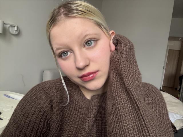 Close-up photo of LenaHoney, fair-skinned with blonde hair, wearing a brown knitted sweater that's wrapped around her head. She has red lipstick, blue eyes, and a slightly flushed face. Background is a simple room.