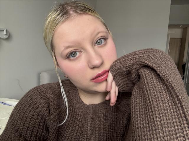 Close-up of LenaHoney, fair-skinned with blonde hair, wearing a brown knit sweater, resting her cheek on her hand, blue eyes, red lips, slight smirk, soft light, casual, slightly provocative.