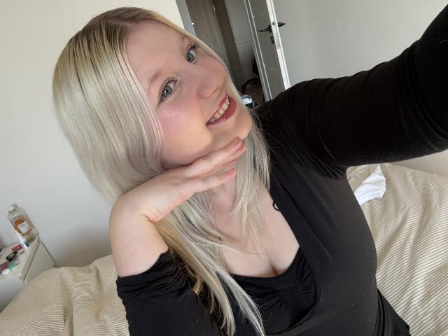 Nahaufnahme von LenaHoney mit blonden Haaren, heller Haut und blauen Augen, die ein schwarzes Oberteil trägt, ihre Hand liegt auf ihrem Hals, und sie blickt auf. Kamera mit einem verführerischen Lächeln. Weiches Licht, Schlafzimmerambiente.