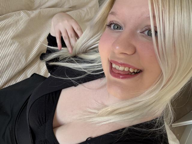 Close-up photo of LenaHoney, blonde, fair-skinned, smiling with red lipstick, lying on a bed. She has blue eyes, black top, and a visible cleavage. Her hand rests on a beige blanket.