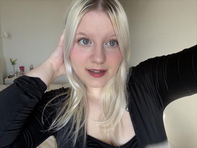 Close-up selfie van LenaHoney met een lichte huid, blauwe ogen en lang blond haar. Ze draagt een zwarte top, haar lippen zijn lichtjes open, en haar rechterhand raakt haar hoofd. De achtergrond is een witte muur. Ongelooflijk aantrekkelijk..