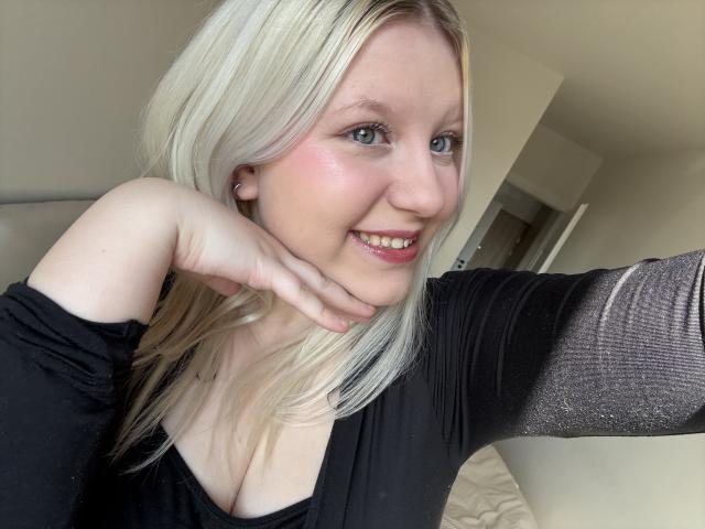 Primo piano di LenaHoney con capelli biondi, pelle chiara e occhi azzurri, che sorride mentre appoggia il mento sulla mano. Indossa una maglia nera con maniche trasparenti, che lascia intravedere il décolleté..