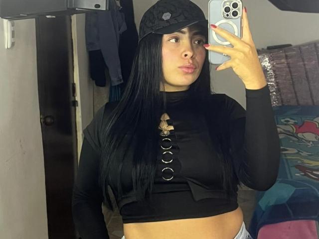 KateWelsin, une femme latina séduisante avec de longs cheveux noirs, porte une casquette noire et un crop top noir moulant avec un laçage sur le devant, et elle prend une autoportrait pris dans un miroir. Elle fait la moue, avec des ongles rouges et un téléphone blanc..