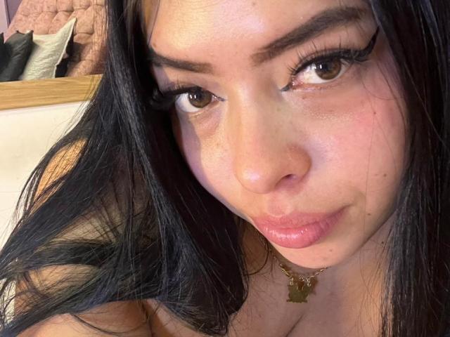 Close-up of KateWelsin, dark hair, brown eyes, thick eyebrows, long eyelashes, glossy pink lips, slight pout, gold cross necklace, bare shoulders, soft lighting, subtle freckles, sexy, intimate, direct gaze.