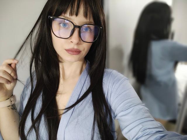 Photographie de NoemiLou, une femme blanche avec de longs cheveux noirs et des yeux bleus, portant de grandes lunettes à monture noire et une tenue claire. Chemise bleue.
son reflet en arrière-plan la montre de dos. Elle tient ses cheveux avec une expression légèrement séduisante..