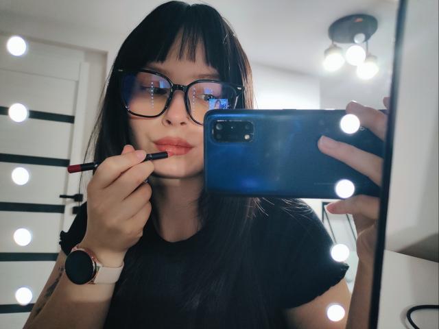 NoemiLou tira uma selfie com um smartphone, segurando um batom vermelho nos lábios. Ela usa óculos pretos, uma camiseta preta e um smartwatch. Lâmpadas brilhantes a cercam. Incrivelmente sexy..