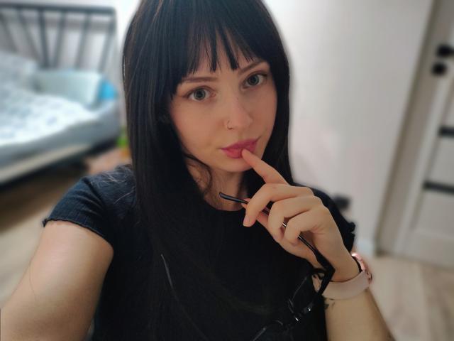 NoemiLou, con cabello negro lacio y flequillo, muerde su dedo de forma seductora. Ella lleva una camisa negra, tiene un piercing en la nariz y un reloj inteligente. El fondo incluye una cama y una puerta desenfocadas..