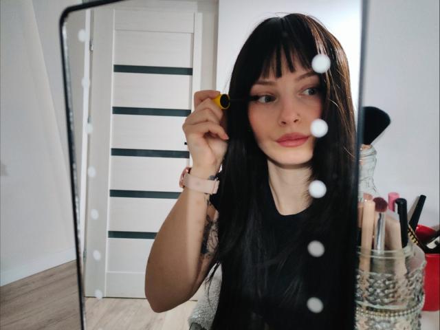 NoemiLou, avec de longs cheveux noirs, appliquant du mascara devant un miroir. Elle tient un applicateur de mascara jaune, et porte un haut noir. Des pinceaux de maquillage sont dans un pot sur la droite. Éclairage lumineux.