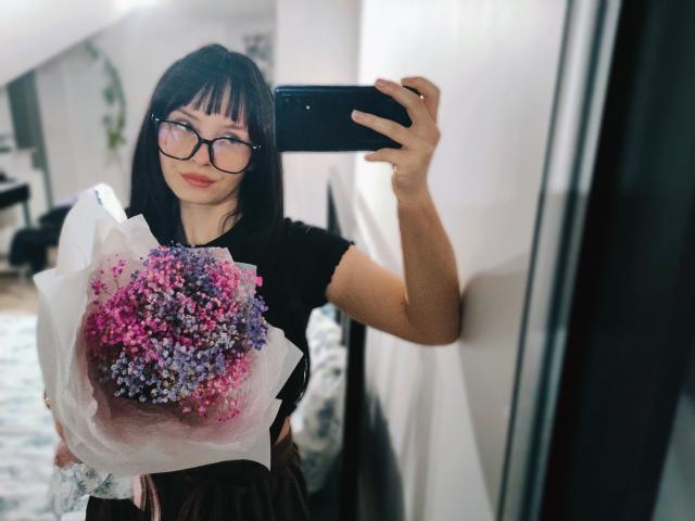NoemiLou, una donna asiatica con capelli neri lisci e occhiali, che tiene un bouquet di fiori rosa e viola, mentre scatta una selfie allo specchio con uno smartphone nero. Sfondo sfocato. Incredibilmente sexy..