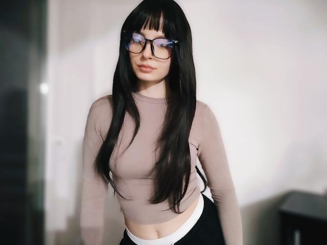 Fotografia de NoemiLou, em pé, vestindo uma blusa cinza transparente de mangas compridas que revela seus seios e calcinha branca. Ela tem cabelos longos e pretos, usa óculos e tem uma expressão sexy e confiante. Fundo claro.