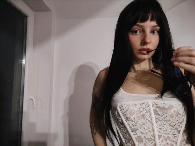 NoemiLou with long black hair, wearing a white lace corset, biting a black strap. Shadow on white wall, dimly lit. Seductive expression, slight smile, focusing on chest area.