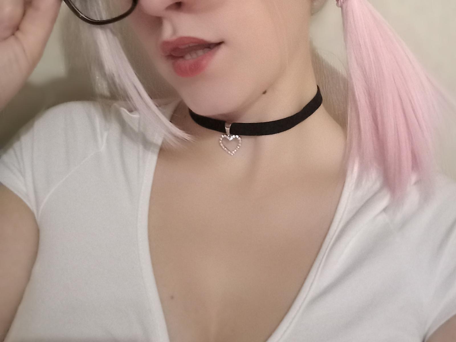 Close-up de AlleksaShshow, com foco em seu pescoço e peito. Ela tem cabelo rosa claro, usa um colar preto com um pingente de coração e uma blusa branca justa que mostra o decote. Seus lábios estão ligeiramente entreabertos..