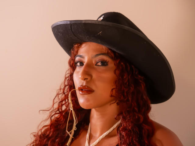 Fotografia de MiaBianchi, usando um chapéu de cowboy preto, com cabelo cacheado ruivo, batom vermelho escuro e um top branco de alças. Sua expressão é sedutora, com uma tensão sexual evidente. Iluminação forte..