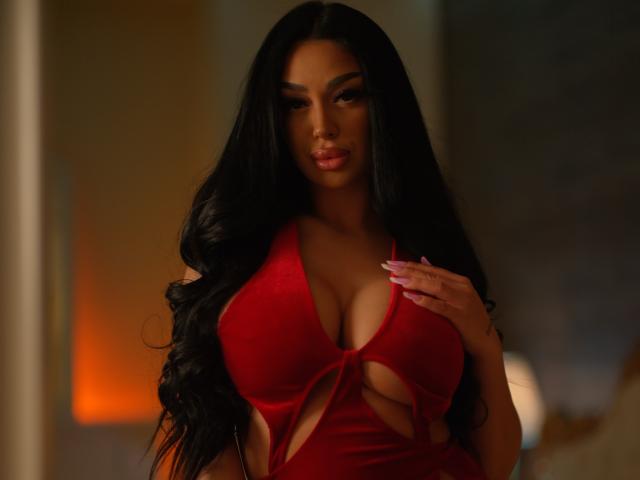 Fotografia de uma mulher curvilínea com cabelos longos e pretos, vestindo um vestido vermelho com recortes, mostrando seios grandes, com a mão peito, expressão sedutora, iluminação fraca, brilho laranja ao fundo.