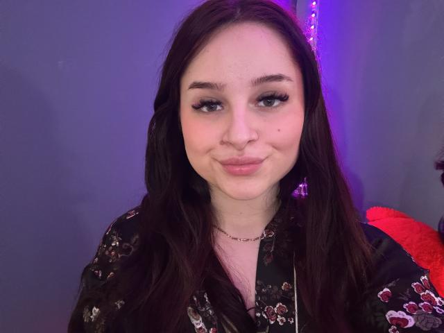 Fotografía de primer plano de LucieRed con piel pálida, cabello largo y castaño oscuro, y maquillaje sutil. Ella está sonriendo ligeramente, y lleva una bata negra con estampado floral. El fondo tiene luces azules y moradas. Qué lindo/a..