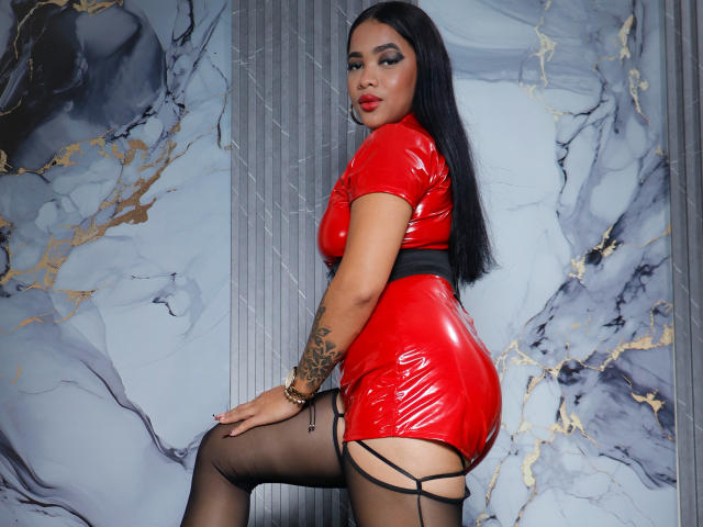 KarlaBella, con un vestido ajustado de látex rojo, una faja negra y medias, mirando de lado con una expresión seductora. Ella tiene el pelo largo y negro, tatuajes en el brazo izquierdo y lápiz labial rojo..