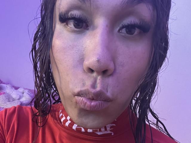 Close-up van GeraldineSuan, een vrouw met nat, donker haar, die haar lippen samendrukt alsof ze een kus geeft. Ze draagt een rood shirt met witte tekst. Haar make-up is zwaar, haar ogen zijn half dicht. Blauw licht.