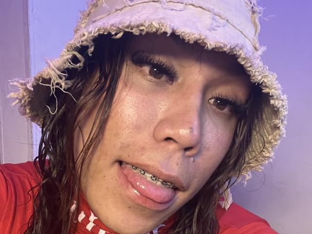 Primo piano di GeraldineSuan, che indossa un cappello di paglia malconcio, con la lingua forata da borchie sporgente, con capelli castani disordinati, rossi camicia e pelle chiara. L'immagine ha un effetto di colore molto saturo, quasi al neon..