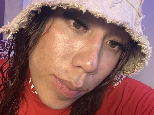 Photo en gros plan de GeraldineSuan, avec des cheveux bruns mouillés, mi-longs, portant un chapeau seau beige usé et une chemise rouge. Son visage est légèrement rougi, ses yeux sont à moitié clos, et ses lèvres sont entrouvertes. Tension sexuelle évidente..