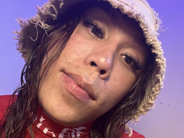 Close-up of GeraldineSuan with messy brown hair, wearing a frayed bucket hat and red shirt. Her skin is slightly freckled, eyes half-closed, lips slightly parted, with a sultry expression. Bright blue sky background.
