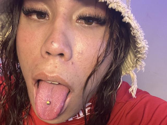 Close-up photo of GeraldineSuan sticking out her tongue with a tongue piercing, brown eyes, long eyelashes, nose piercing, messy brown hair, wearing a straw hat and red shirt. Fucking sexy.