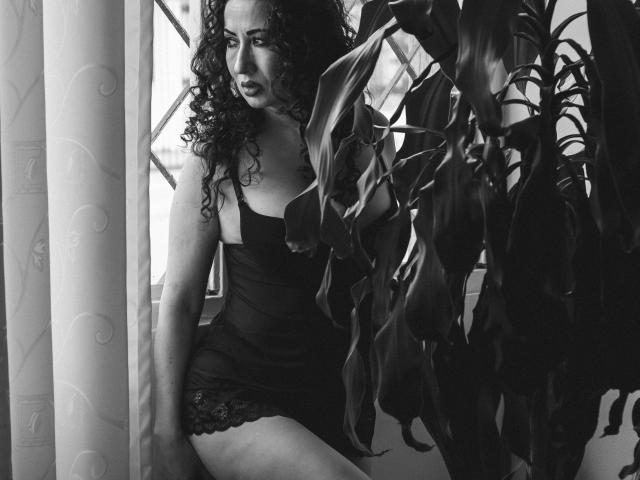 Fotografia in bianco e nero di Samantha Antolin, una donna formosa con lunghi capelli ricci, seduta tra piante alte, che indossa un abito nero aderente in pizzo, con il suo pene visibile attraverso il tessuto, sguardo intenso.