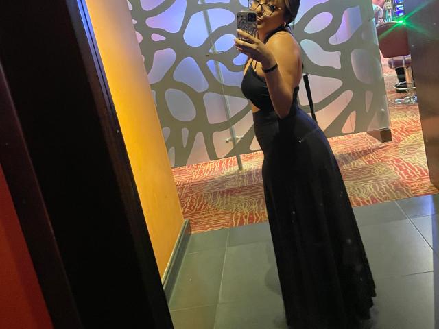 DulceLiz, in a tight black dress, stands in a mirror shot, holding a phone. She's sexy with a curvy figure, dark skin, and short hair. Background has patterned walls.