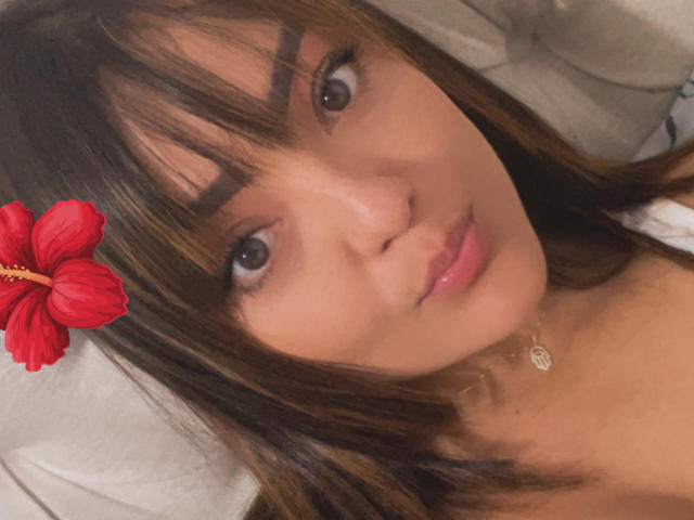 Selfie de DulceLiz en primer plano, con piel de color marrón medio, cabello castaño lacio, flequillo y una flor de hibisco roja en el cabello. Ella lleva un collar de oro y mira a la cámara con una ligera sonrisa..